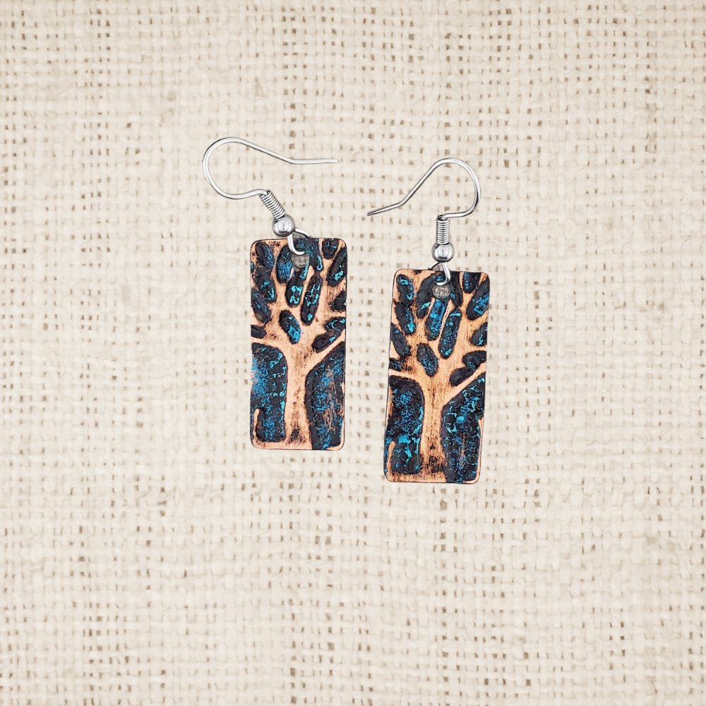 Pair of vertical rectangular copper earrings with embossed tree design and turquoise patina on a textured beige background