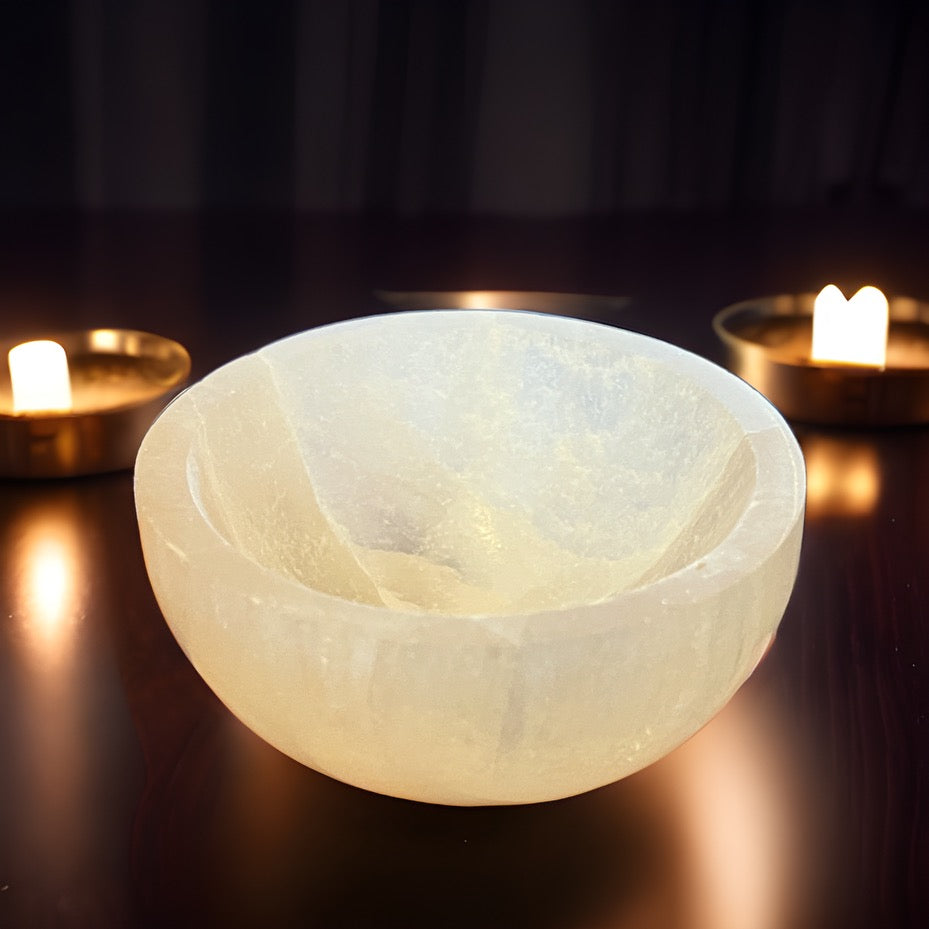 Carved white selenite bowl sitting on a reflective black counter with candle flame behind it.
