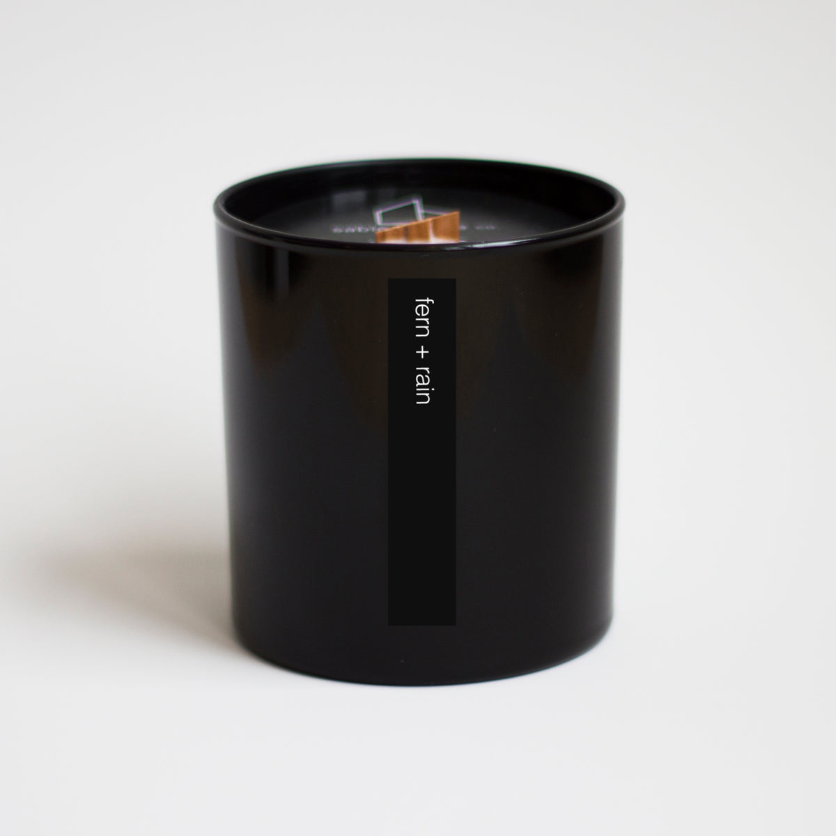 Candle with wood wick inside black canister with the words &quot;fern + rain&quot; in white lower case letters.