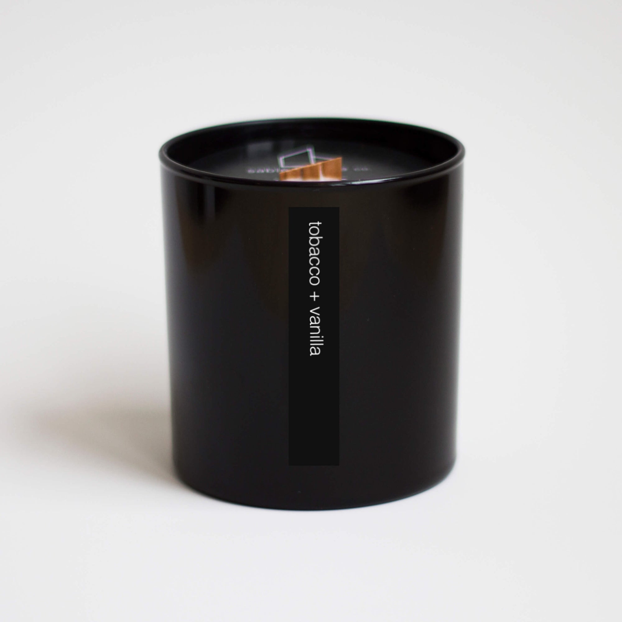 Tall black container holding beige candle with wood wick. Words on outside read "tobacco + vanilla".