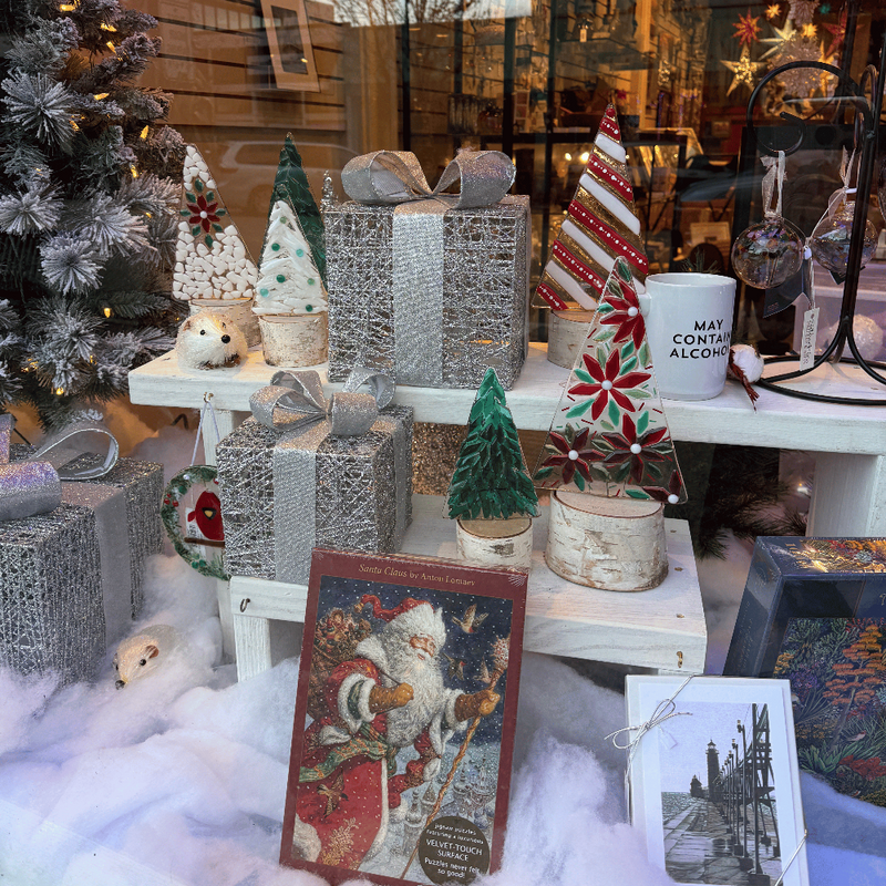 A festive Christmas-themed display in a shop window featuring wrapped gift boxes, miniature Christmas trees, a Santa Claus card, a mug reading 'MAY COMFORT ALCOHOL', and holiday decorations arranged on white shelves with faux white faux-fur styling.