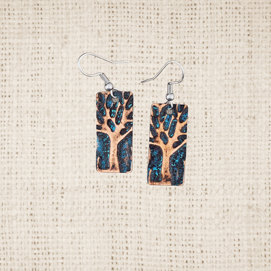 Pair of vertical rectangular copper earrings with embossed tree design and turquoise patina on a textured beige background
