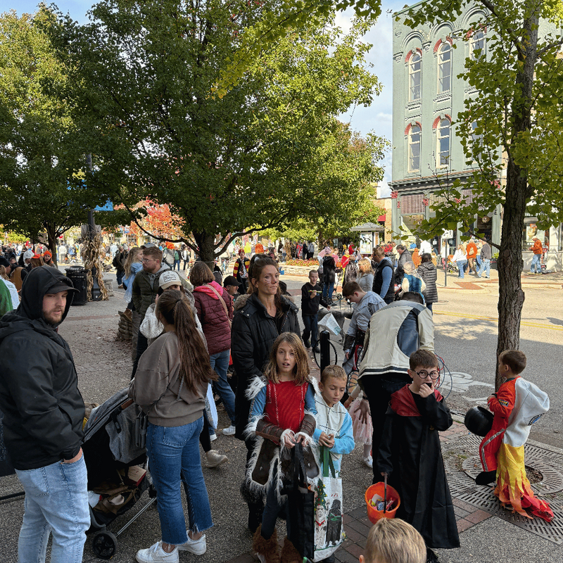 A busy outdoor street scene with a crowd of people, trees, and a historic building in the background. Several children are wearing costumes, including a dragon costume on the right and a knight costume in the foreground.