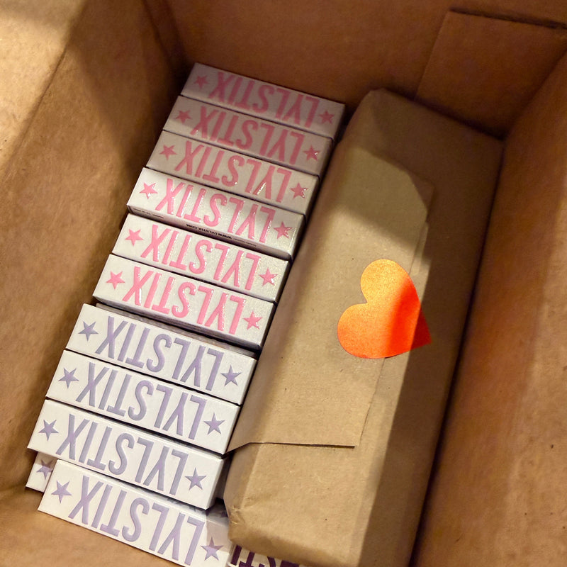 A cardboard box containing multiple white rectangular items printed with the brand name 'LYLSTIX' in pink, purple, and blue, alongside a brown paper-wrapped package with a small orange heart sticker.