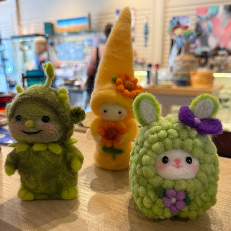 Three small felt plush toys displayed on a wooden table in a shop setting. The left toy is olive green with antennae and a smiling face.