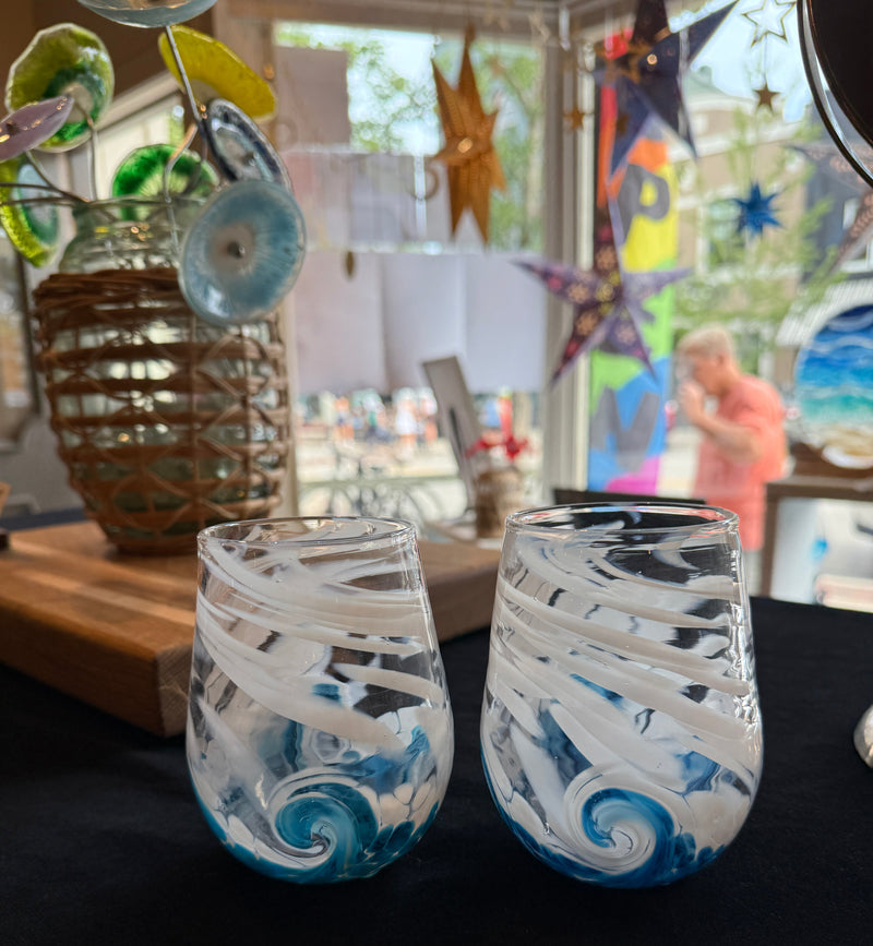 Two hand-blown stemless wine glasses with white and blue swirl patterns, displayed on a black tablecloth in a shop setting.