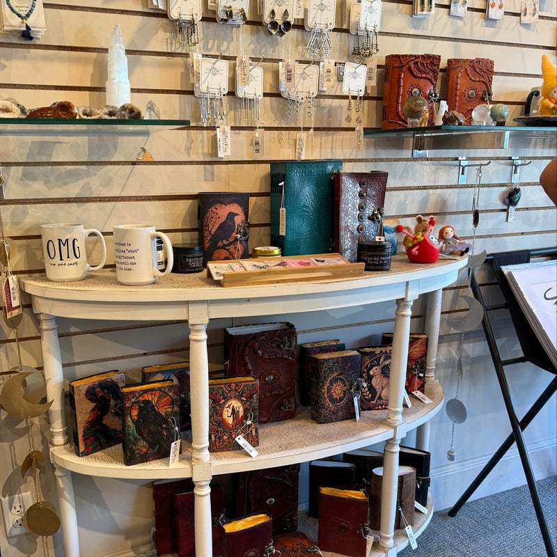 A rustic white wooden three-tier display shelf in a shop, filled with various decorative items including mugs, books, and small boxes. The background features a light wood slatwall with many hanging earrings.