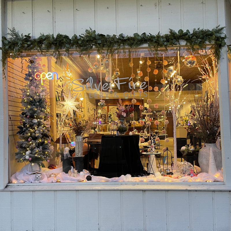 SilverFire Gallery & Gifts' window display decorated for the holidays, featuring a Christmas tree, warm string lights, floral arrangements, and green garlands across the top of the window.