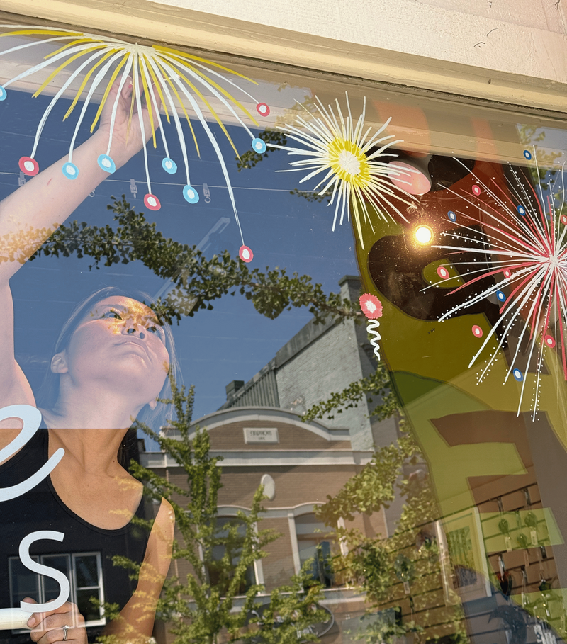 A storefront window display featuring a large printed mural of a woman in a black tank top, decorated with colorful hanging balloon-like lights and white fireworks graphics. Strong reflections of a street scene with trees, buildings, and sky are visible on the glass.