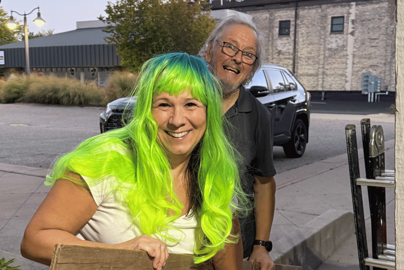 A woman with bright neon green hair smiles while holding a large wooden board outdoors, with a man standing behind her carrying another board. Cardboard boxes and a ladder are visible on the ground beside them.