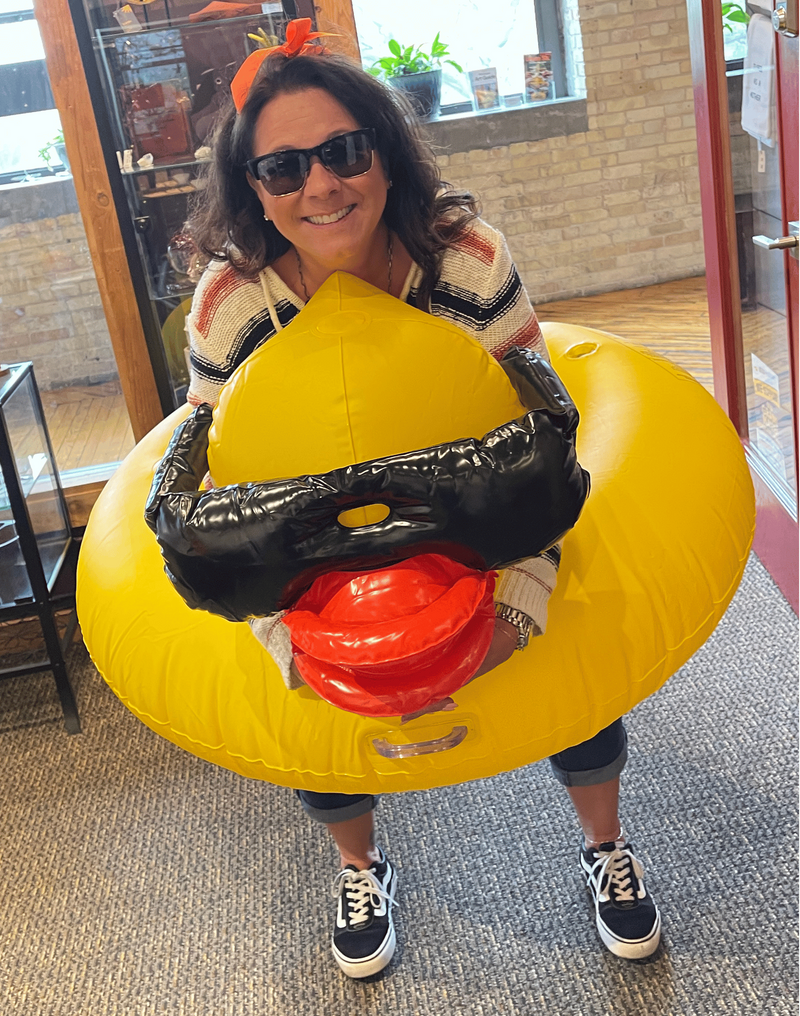 A woman wearing sunglasses and a striped shirt stands indoors holding a large inflatable yellow duck costume with black beak details and a red mouth.
