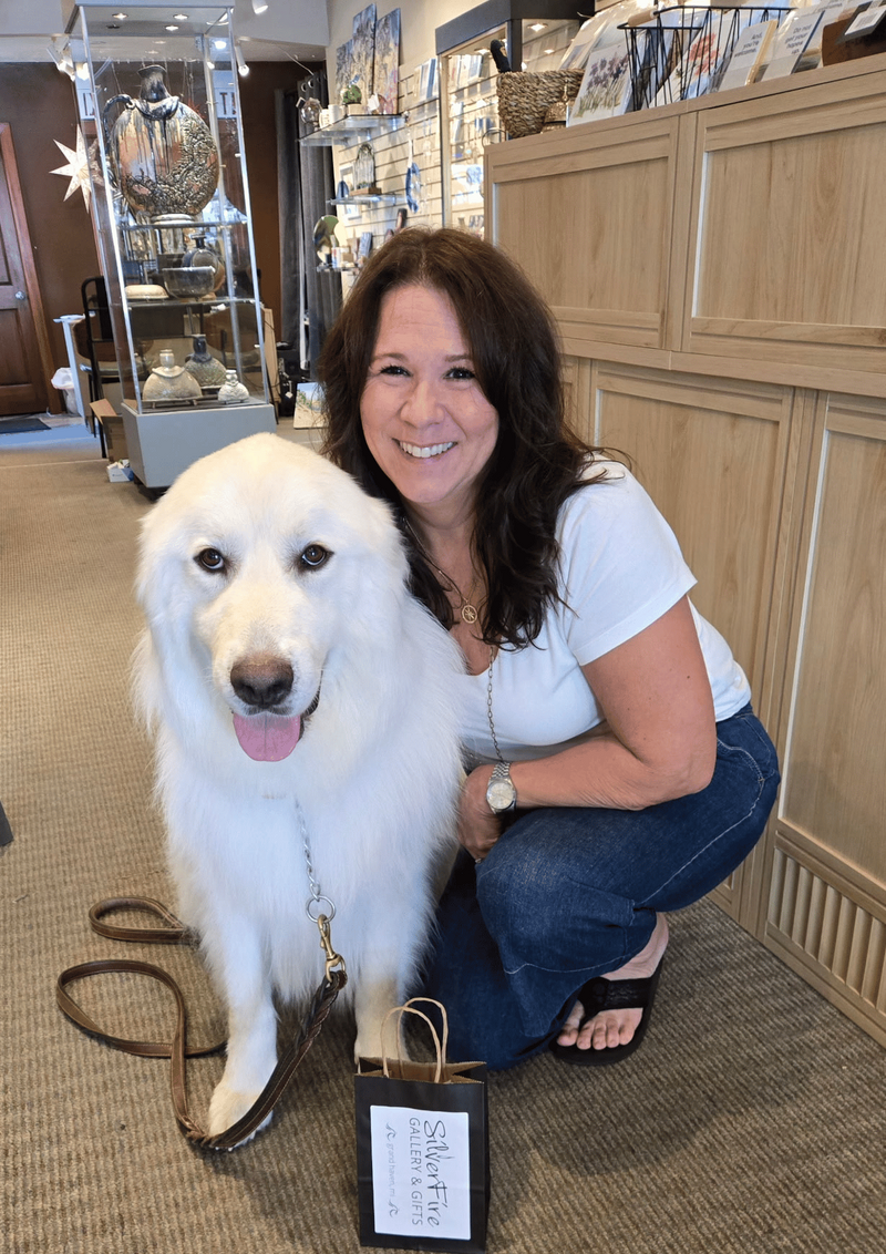 A smiling woman crouches beside a large white dog in a retail store, holding a small gift bag with a tag in front of them. The store has light wood cabinets and shelving in the background.