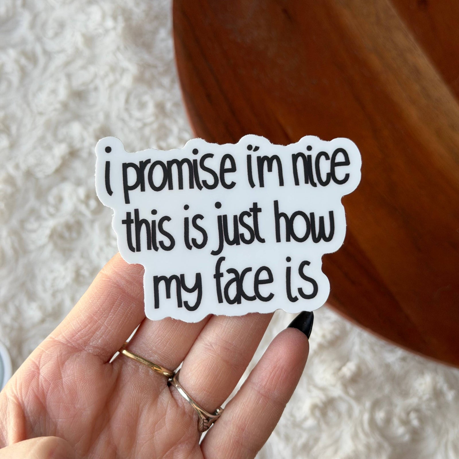 Hand holding a sticker with text 'I promise I'm nice this is just how my face is' against a neutral background.