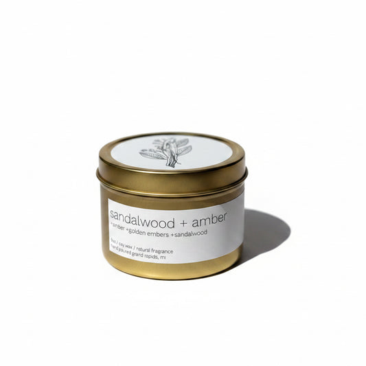 A gold metal tin candle with the label 'Sandalwood + Amber' in minimalist font, placed on a white surface with shadow patterns.