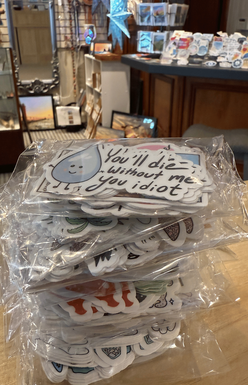 A stack of clear plastic-wrapped sticker packs on a wooden table inside a retail store. The top sticker reads 'You'll die without me you idiot' with a blue water drop smiling face illustration.