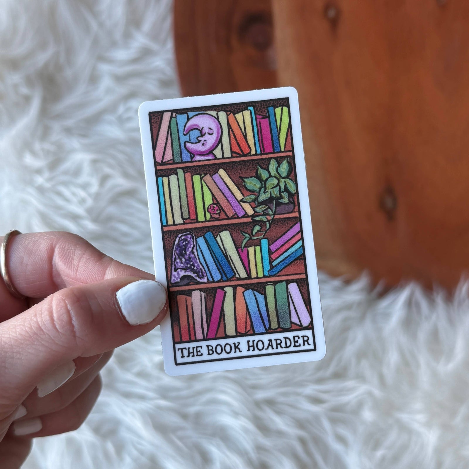 Card titled 'The Book Hoarder' held by a hand on a white fluffy surface with wooden background