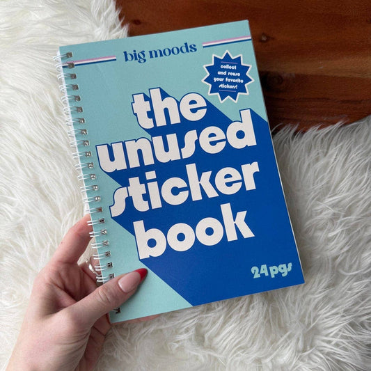 Hand holding 'the unused sticker book' by big moods on a fluffy white surface