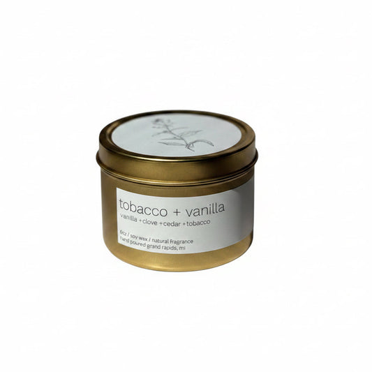 A gold tin candle with the label 'Tobacco + Vanilla' placed on a white surface.