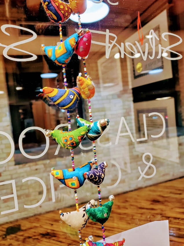 A colorful hanging ornament made of small bird-shaped beads and tassels, photographed in front of a glass window with reflections and white script text.
