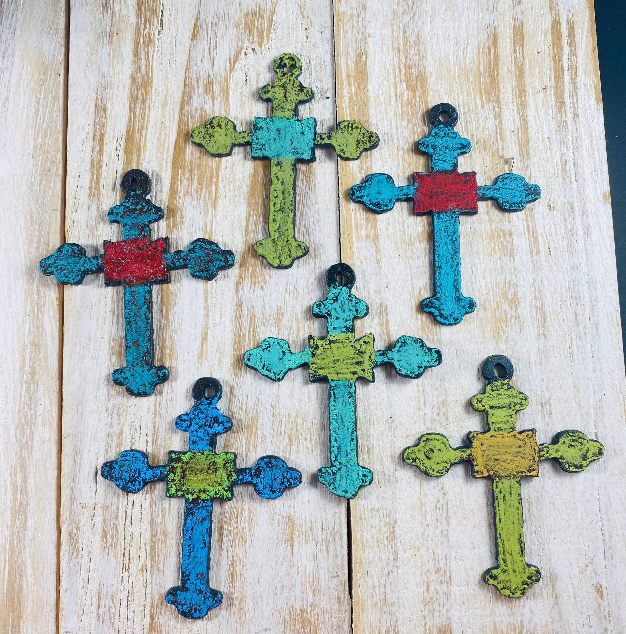 White-washed wood table holds six metal stylized rustic crosses painted in bright color combinations including turquoise and red, lime and turquoise, light turquoise and red, light teal and lime, blue and lime, lime and yellow. 