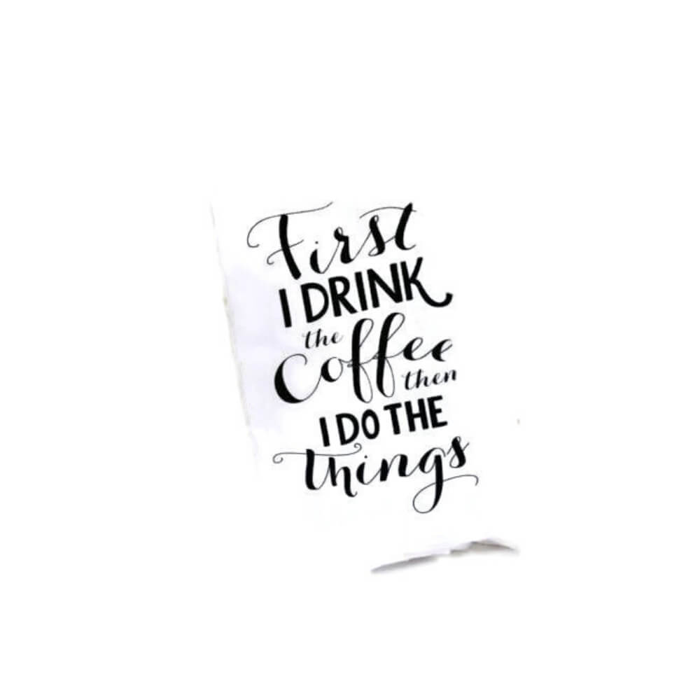 White cotton tea towel with assorted black script and the quote &quot;First I drink the coffee, then I do the things&quot;.