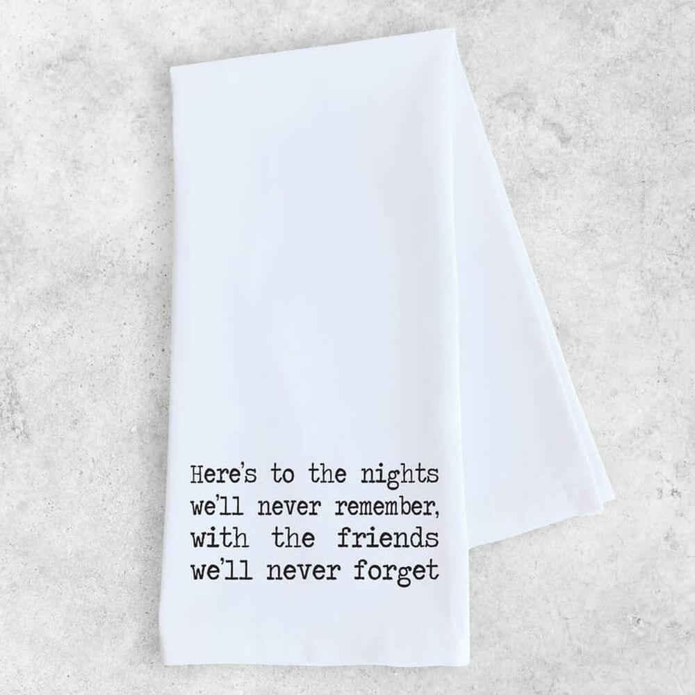 White cotton kitchen towel with typewriter font in black  which reads &quot;Here&#39;s to the nights we&#39;ll never remember, with the friends we&#39;ll never forget.&quot;