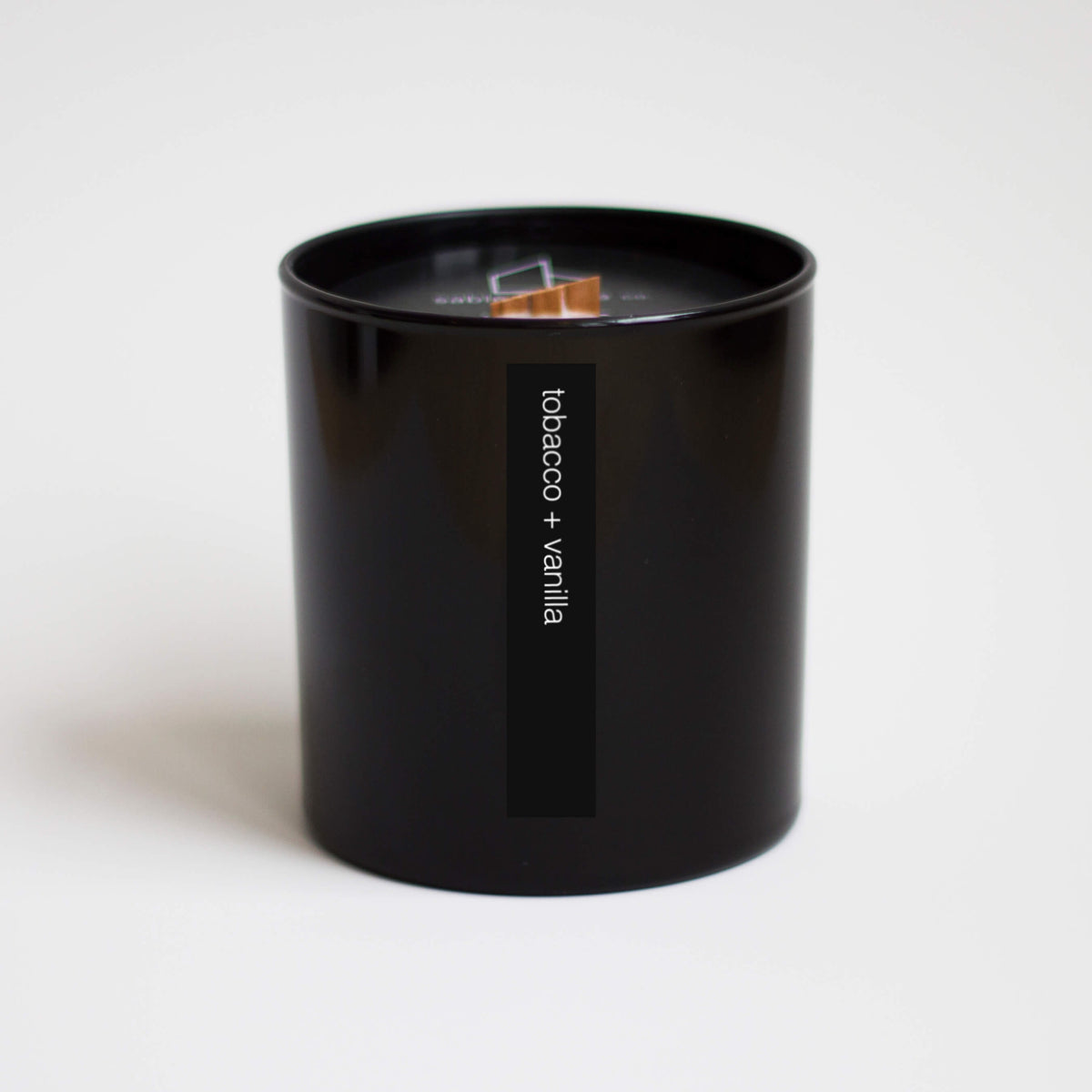 Tall black container holding beige candle with wood wick. Words on outside read &quot;tobacco + vanilla&quot;.