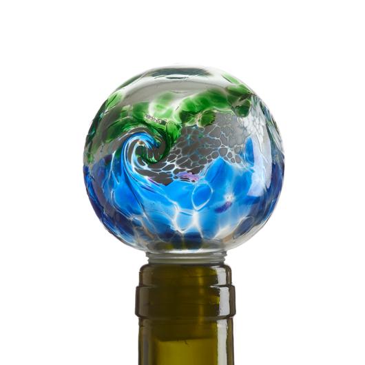 Blown glass globe wine stopper with spatterings of transparent green on the top half and transparent blue on the bottom. The two colors meet in a swirl of white and gray that looks like a wave. Pictured in the neck of a green wine bottle.