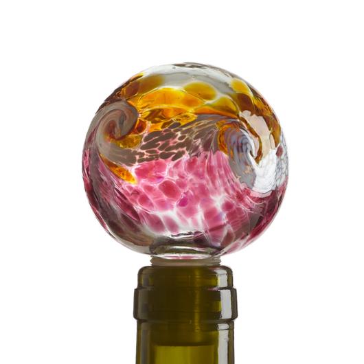 Blown glass globe wine stopper with spatterings of transparent yellow on the top half and transparent pink on the bottom. The two colors meet in a swirl of white and gray that looks like a wave. Pictured in the neck of a green wine bottle.